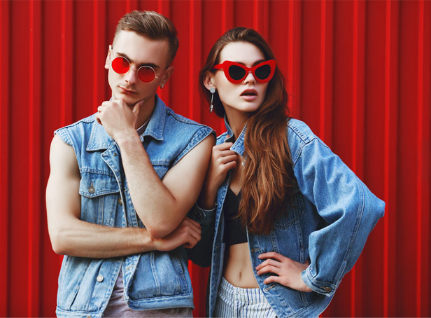 young man and woman wearing denim jackets and red sunglasses against red metal background