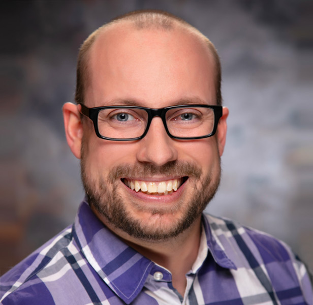 Aaron Bradley smiling, wearing glasses and purple plaid button-down shirt